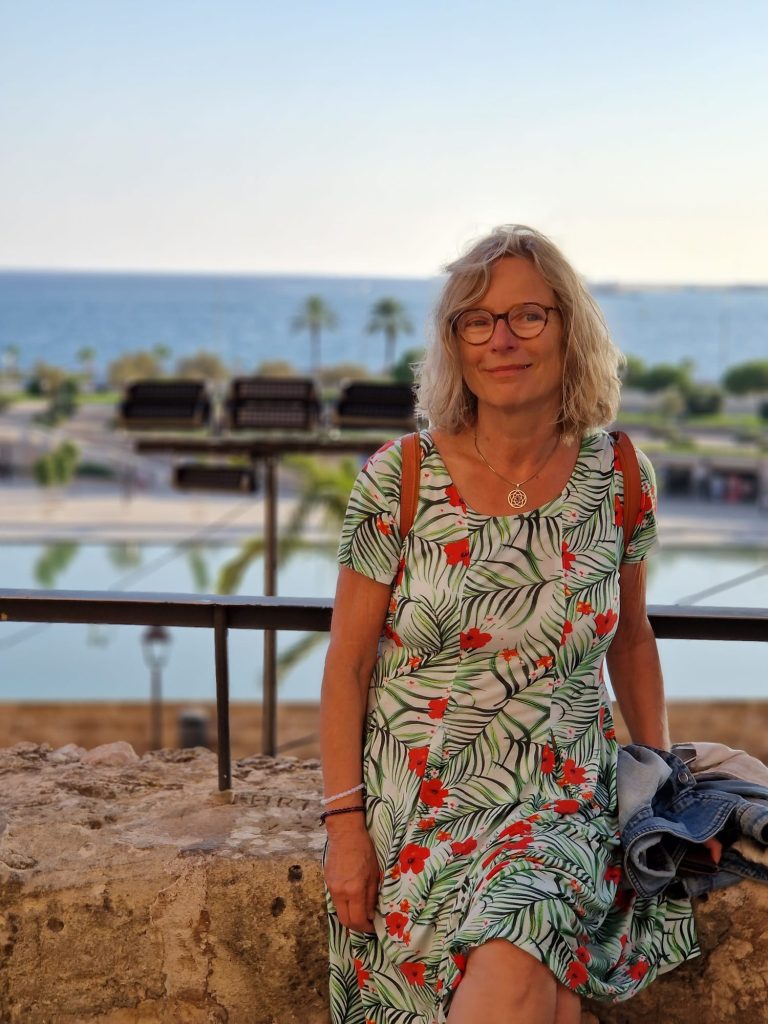 Eine Frau mit Blumenkleid lächelnd, im Hintergrund das Meer und Palmen